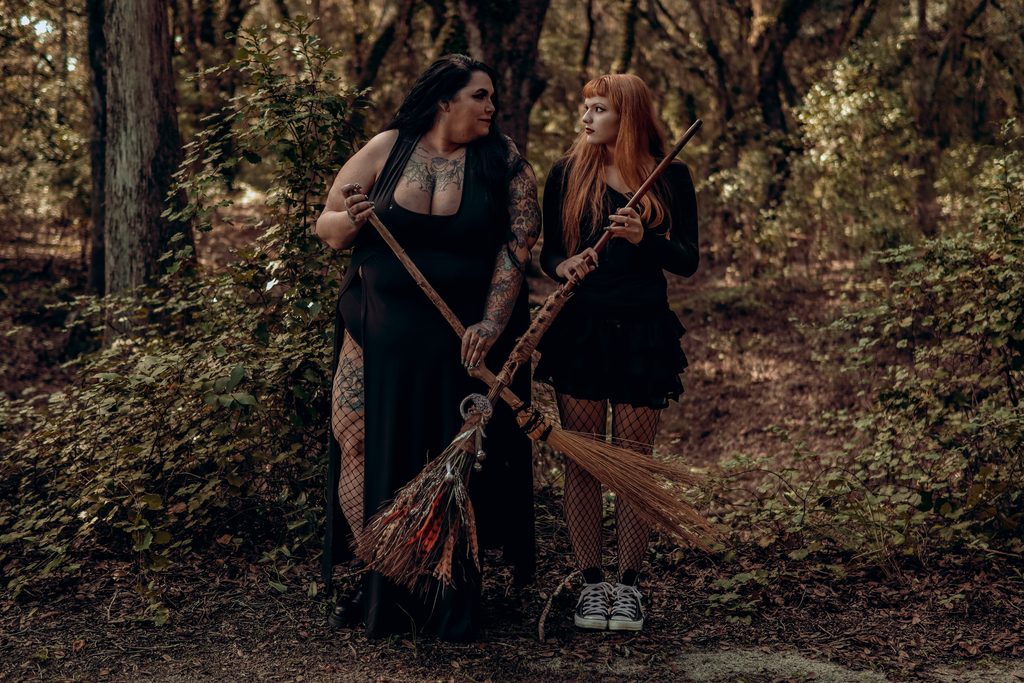 Two dark fantasy models standing together in a sunlit forest, one wearing a black gown and the other holding a woven broom, captured in a witchy Central Florida portrait. witchy fine art portraits