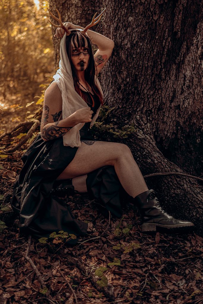 Dark fantasy witch seated amidst forest roots wearing ornate headpiece and dark boots, witchy portrait in Central Florida